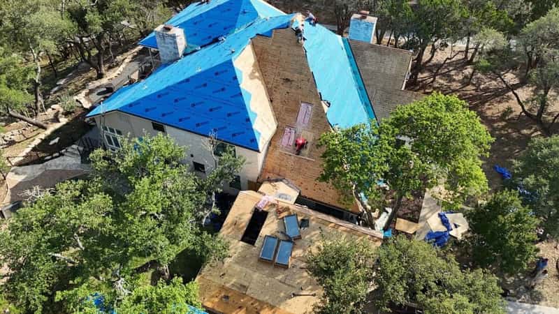 Aerial view of a large home mid-reroof with blue synthetic underlayment and distant crew members