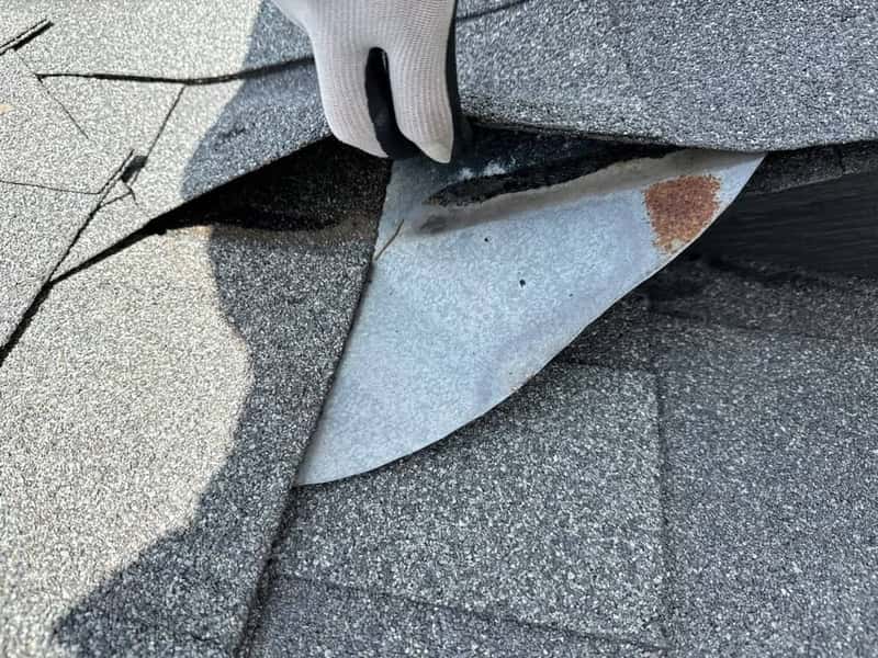 Gloved hand lifting a rusted metal flashing piece tucked under shingles