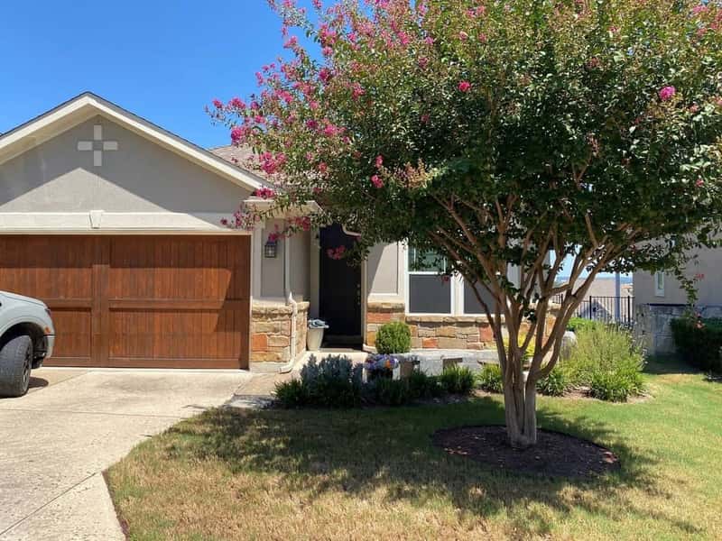 Single-story tan stucco home with wood garage door and a crepe myrtle tree blooming pink