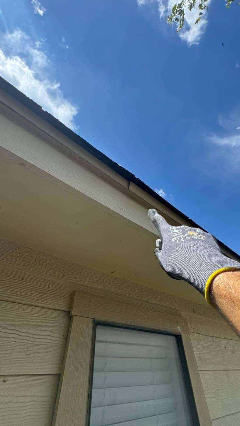 Gloved hand pointing out fascia and eave trim beneath a shingle roofline