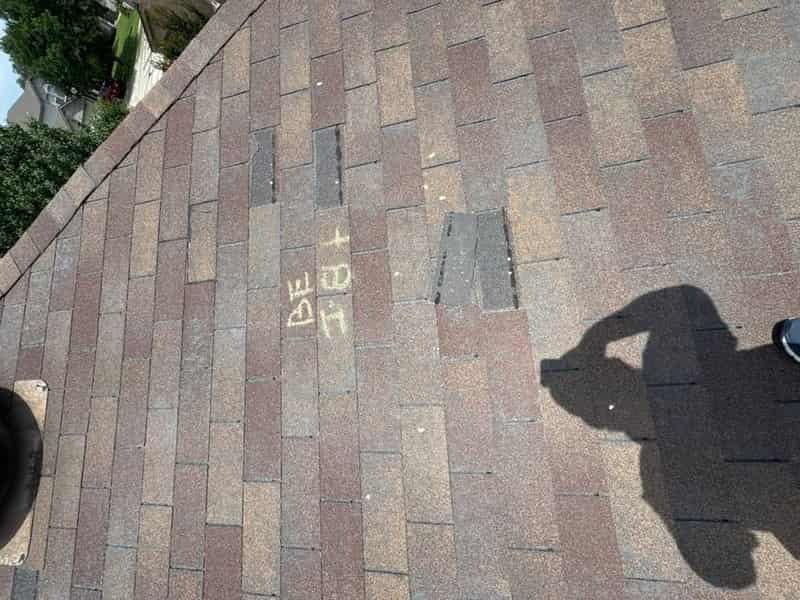 Shingle roof marked with chalk hail-inspection lettering and a worker's shadow cast across it