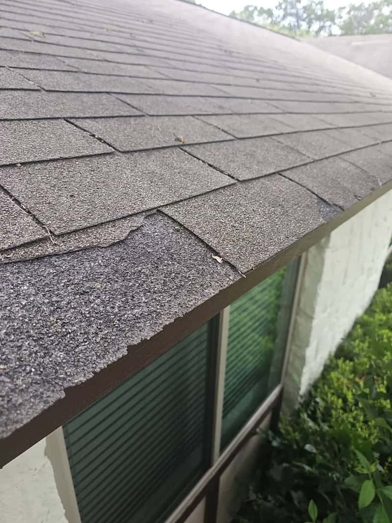 Close-up of curling and cracked shingles hanging over a roof edge above a window