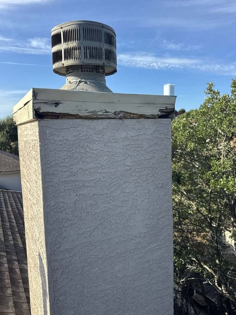 Chimney cap with rotted wood trim and a metal spark arrestor against a blue sky